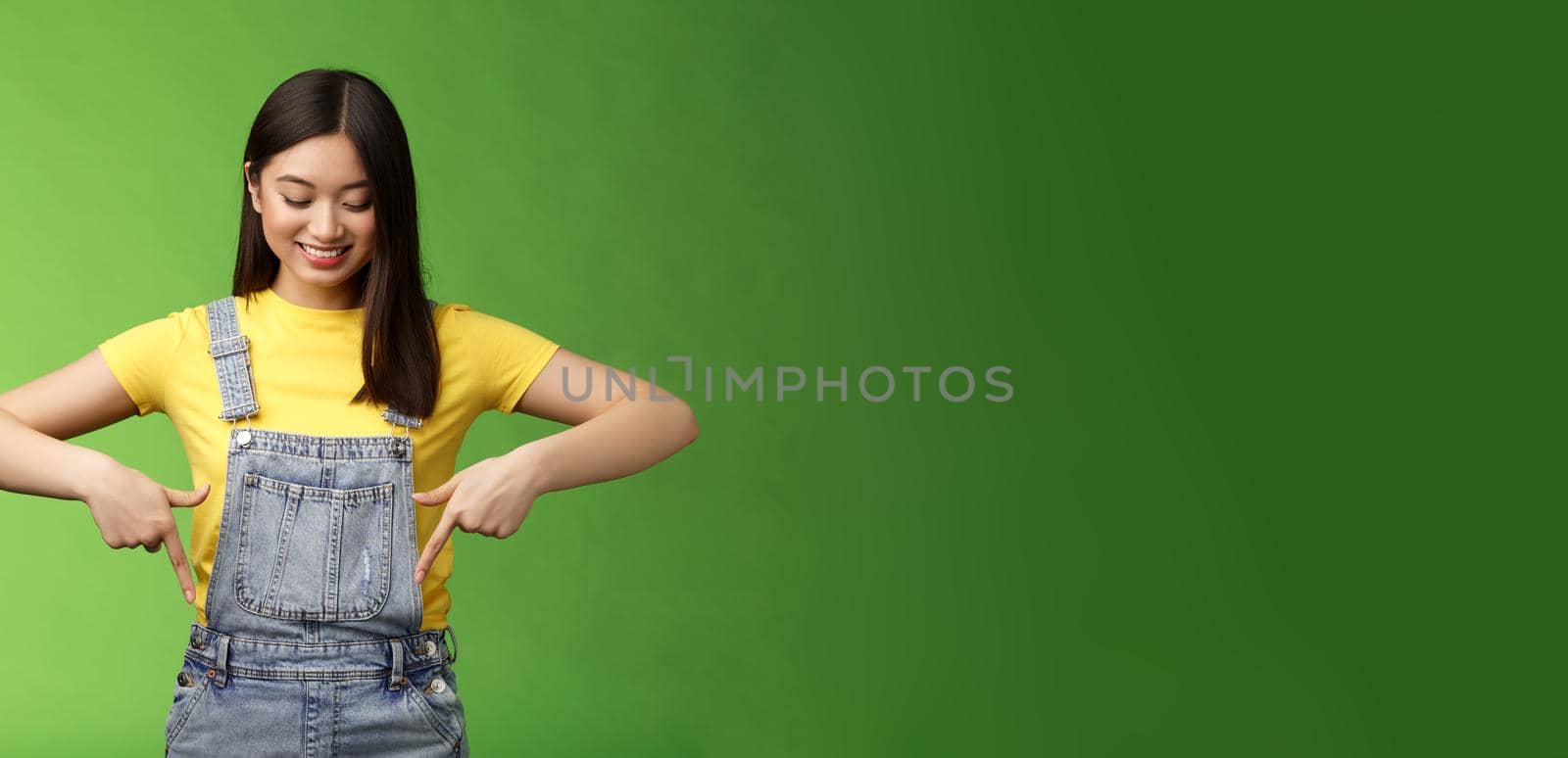 Studio shot easygoing cute asian female introduce new promo, pointing looking down amused pleasantly smiling, gladly show interesting thing, stand green background happy excited. Copy space