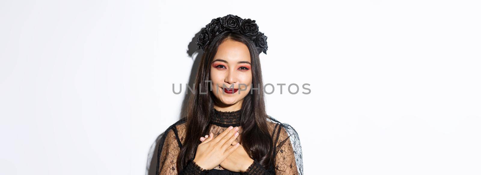 Close-up of thankful smiling asian woman looking grateful with hands over chest, standing in witch costume over white background.