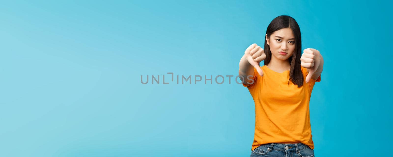 Upset gloomy asian girl with dark haircut frowning sulking sad, disappointed, show thumbs-down grimacing, give negative opinion, judging bad uninteresting movie, stand blue background by Benzoix