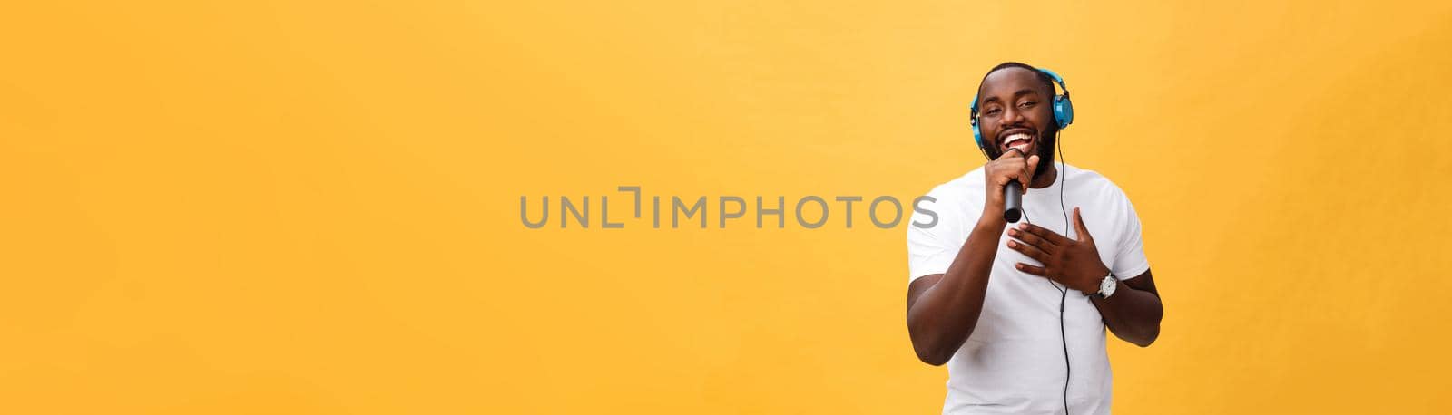 Portrait of cheerful positive chic. handsome african man holding microphone and having headphones on head listening music singing song enjoying weekend vacation isolated on yellow background.