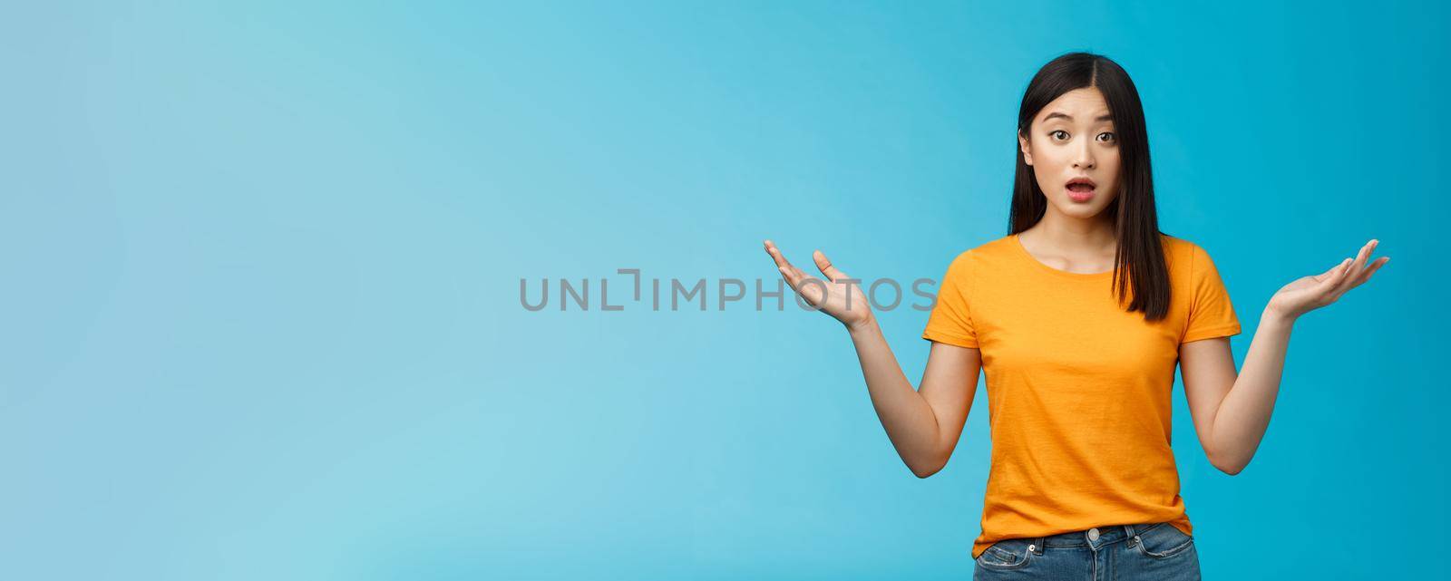 Questioned unaware young asian girl with dark short haircut shrugging hands spread sideways, look confused open mouth uncertain, being clueless, stand blue background wear yellow t-shirt by Benzoix