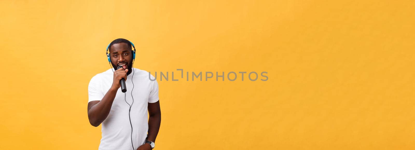 Portrait of cheerful positive chic. handsome african man holding microphone and having headphones on head listening music singing song enjoying weekend vacation isolated on yellow background.