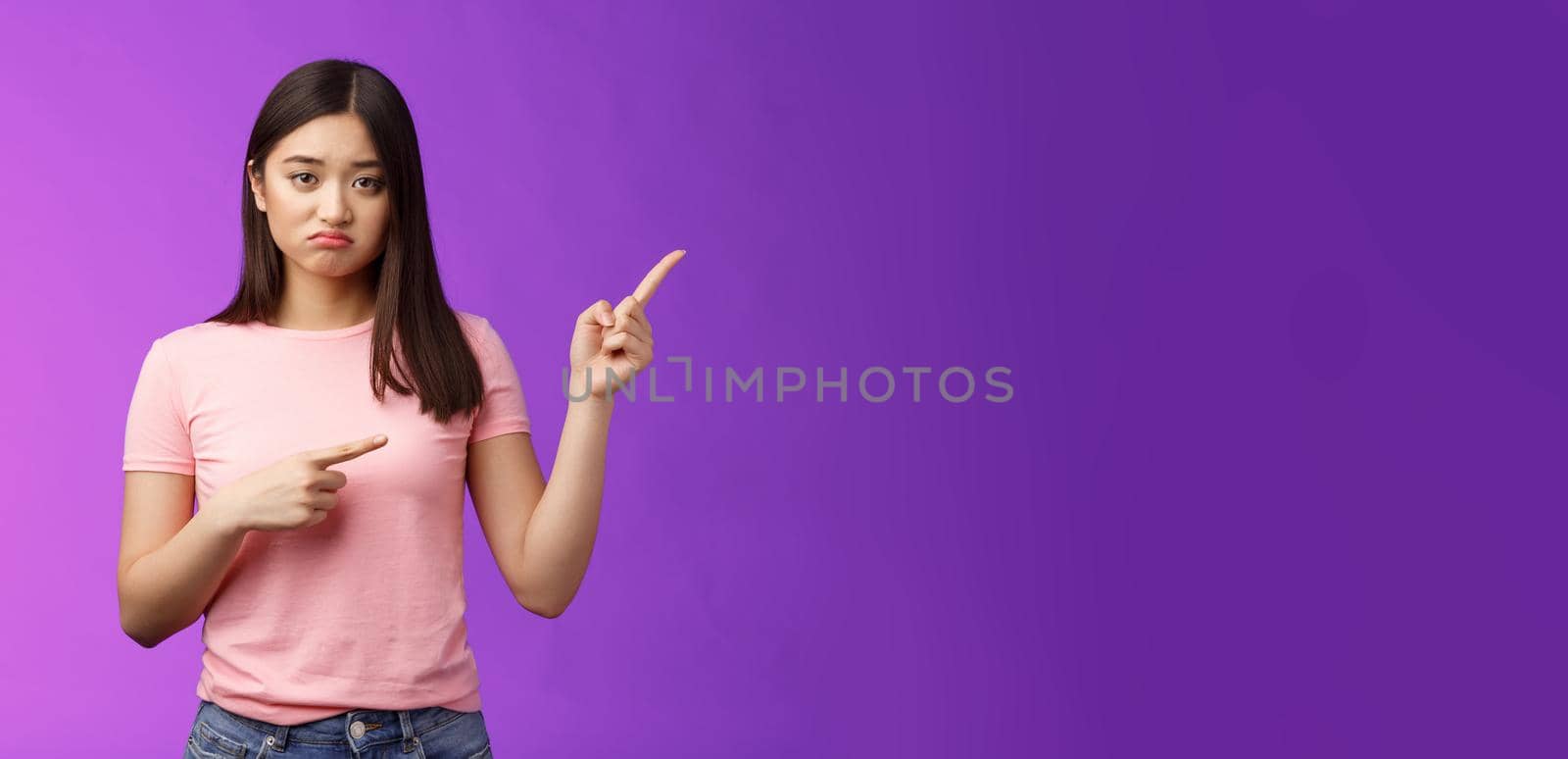 Upset unlucky cute sulking asian girl losing competition, pouting grimacing unhappy, pointing right, indicating fingers sideways with regret and sorrow, feel pitty herself, stand purple background.