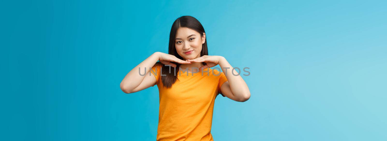 Sweet lovely tender asian girl brunette, tilt head smiling show clean pure face hold palms under jawline satisfied after applying skincare product stand blue background wear yellow t-shirt.