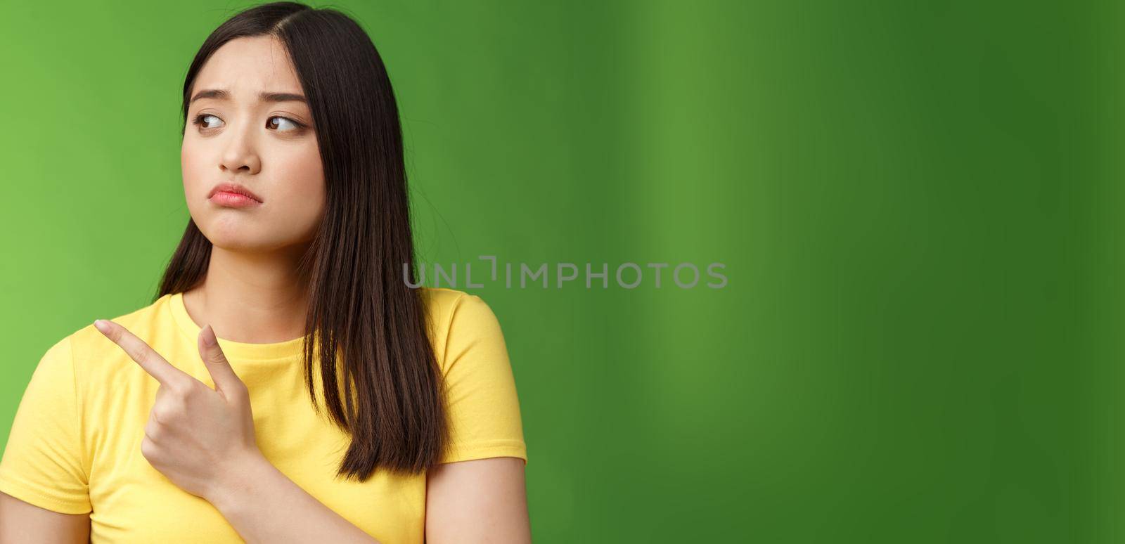 Gloomy upset lonely asian girl jealous sister pointing turn head left, sulking sighing sorrow distress, stand green background unhappy, feel unlucky and disappointed, regret not going concert.