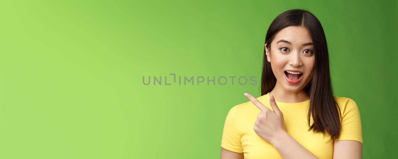 Enthusiastic impressed cute asian brunette describing new haircare product features amused and amazed, lively talking poining left astonished, have interesting discussion, stand green background.