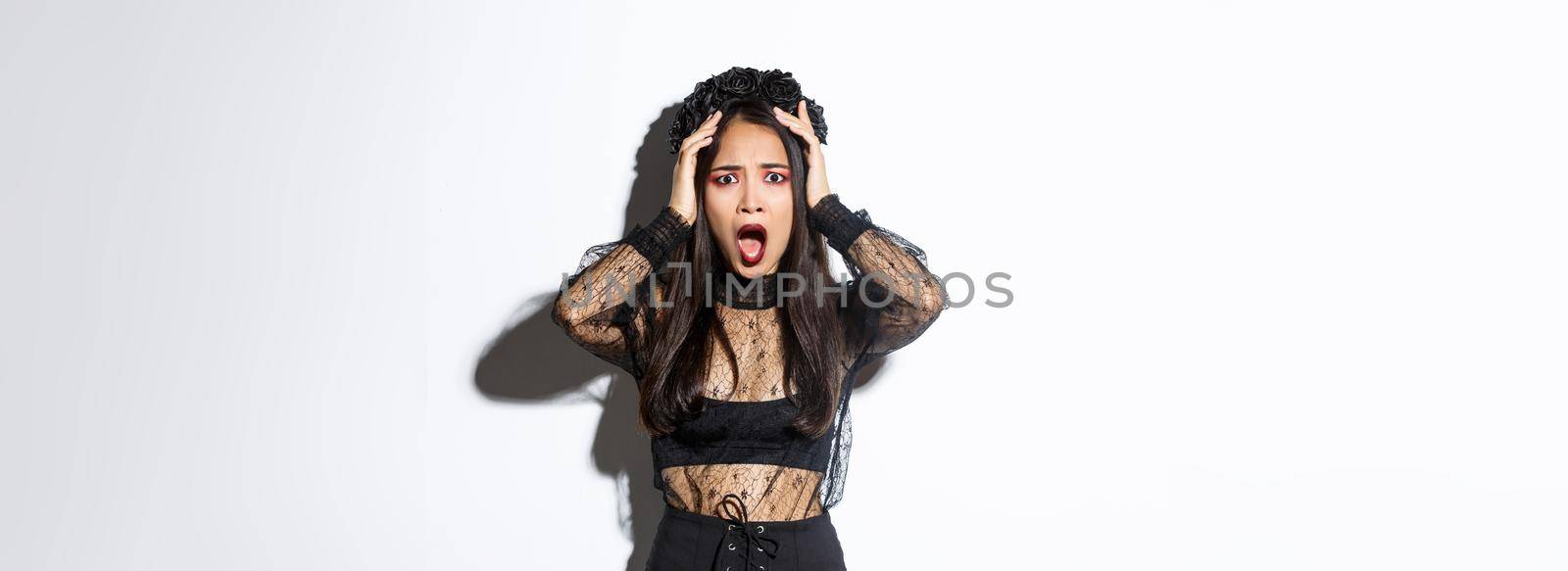 Image of horrified and shocked asian woman in gothic lace dress and wreath looking ambushed, wearing halloween costume, gasping and looking concerned, standing over white background.