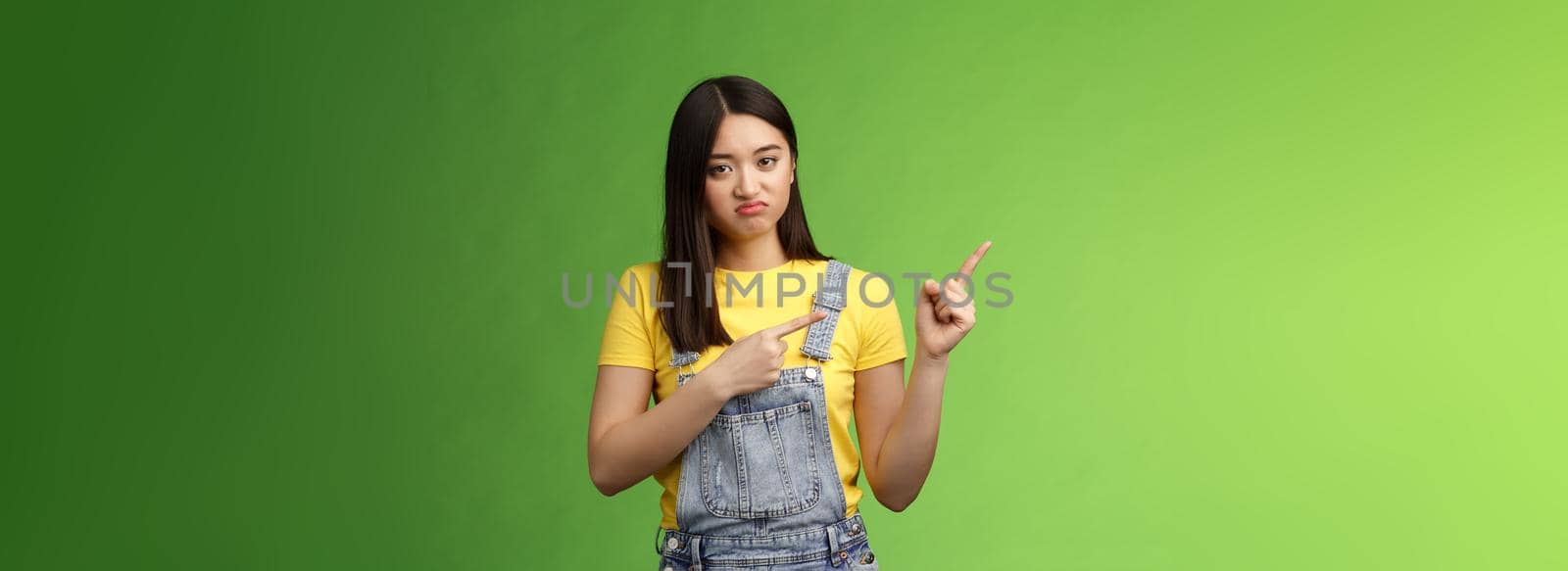Disappointed uninterested asian gloomy girl pointing fingers left, grimacing sulking displeased, unwilling take part event, stare reluctant unimpressed, pose green background. Copy space