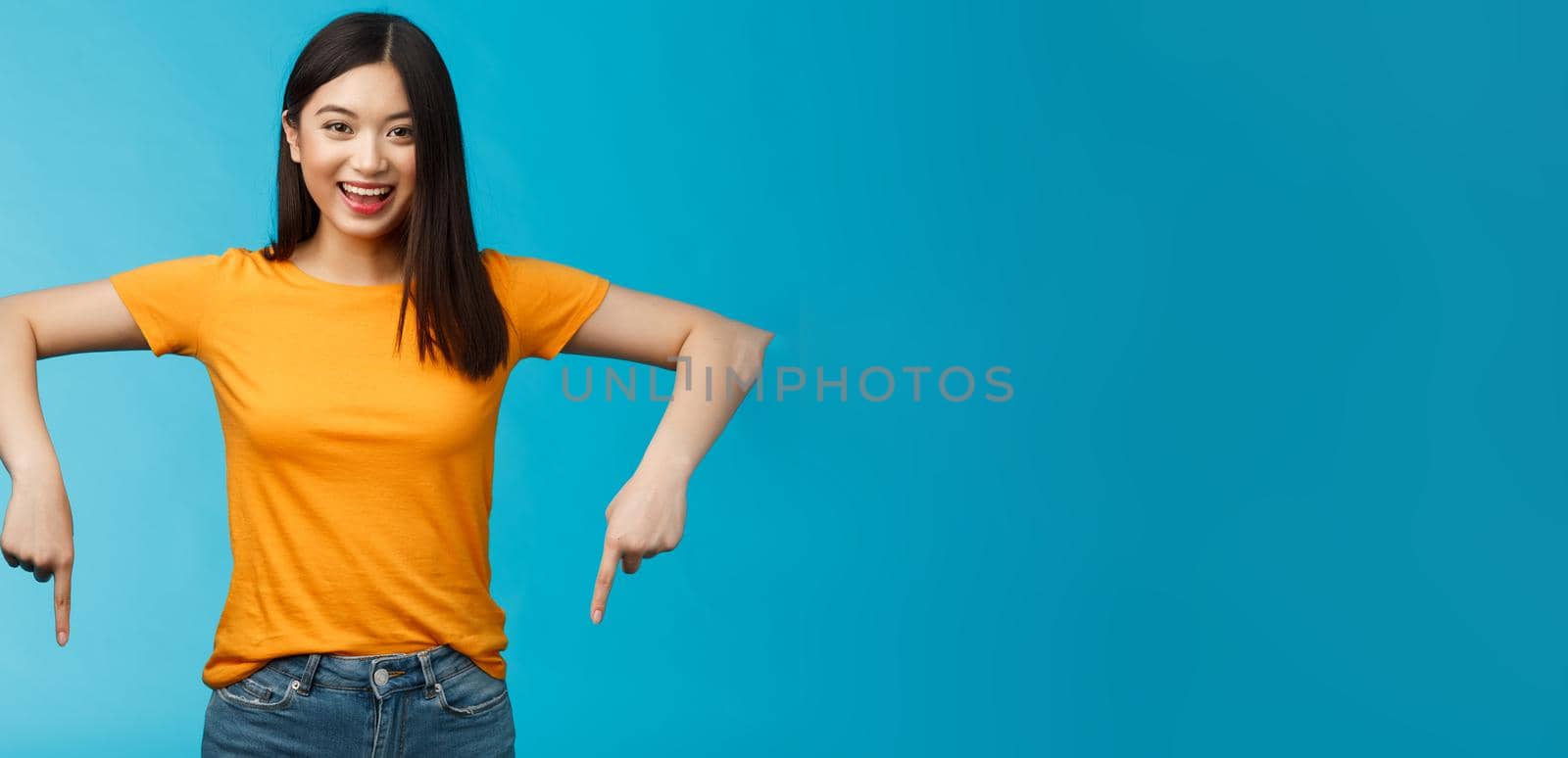 Enthusiastic pleasant asian blong modern female student inviting you visit awesome event pointing down indicating bottom advertisement, stand blue background smiling broadly look happily camera.