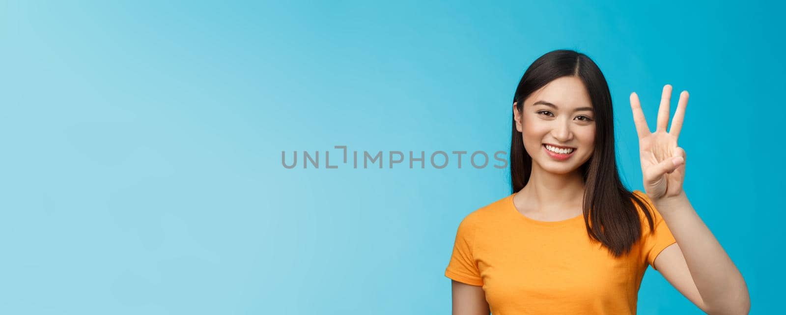 Friendly enthusiastic tender asian woman making reservation for three, number third, smiling broadly, stand blue background joyfully, posing near blue background in yellow t-shirt.