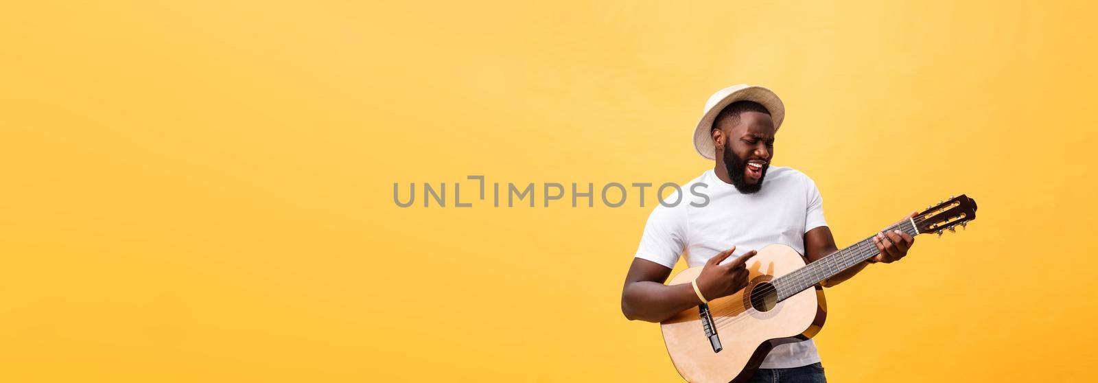 Muscular black man playing guitar, wearing jeans and white tank-top. Isolate over yellow background