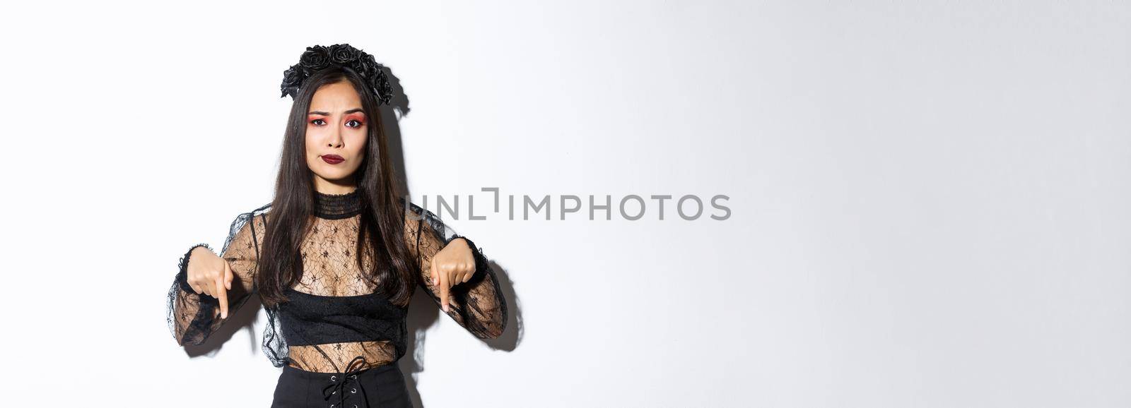 Concerned an disappointed asian woman in black lace dress and wreath smirk skeptical while pointing fingers down at something bad, complaining over white background by Benzoix