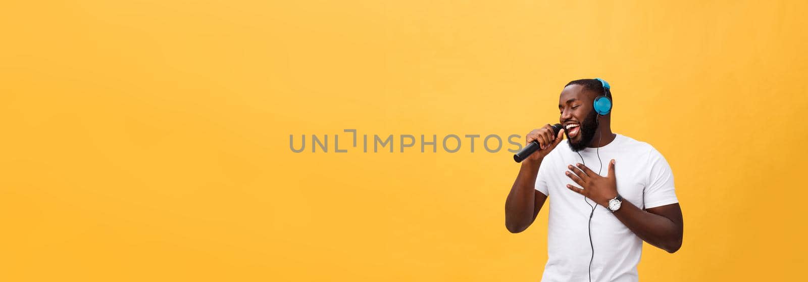 Portrait of cheerful positive chic handsome african man holding microphone and having headphones on head listening music singing song enjoying weekend vacation isolated on yellow background by Benzoix