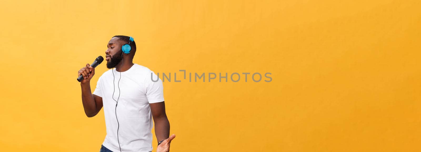 Portrait of cheerful positive chic. handsome african man holding microphone and having headphones on head listening music singing song enjoying weekend vacation isolated on yellow background.