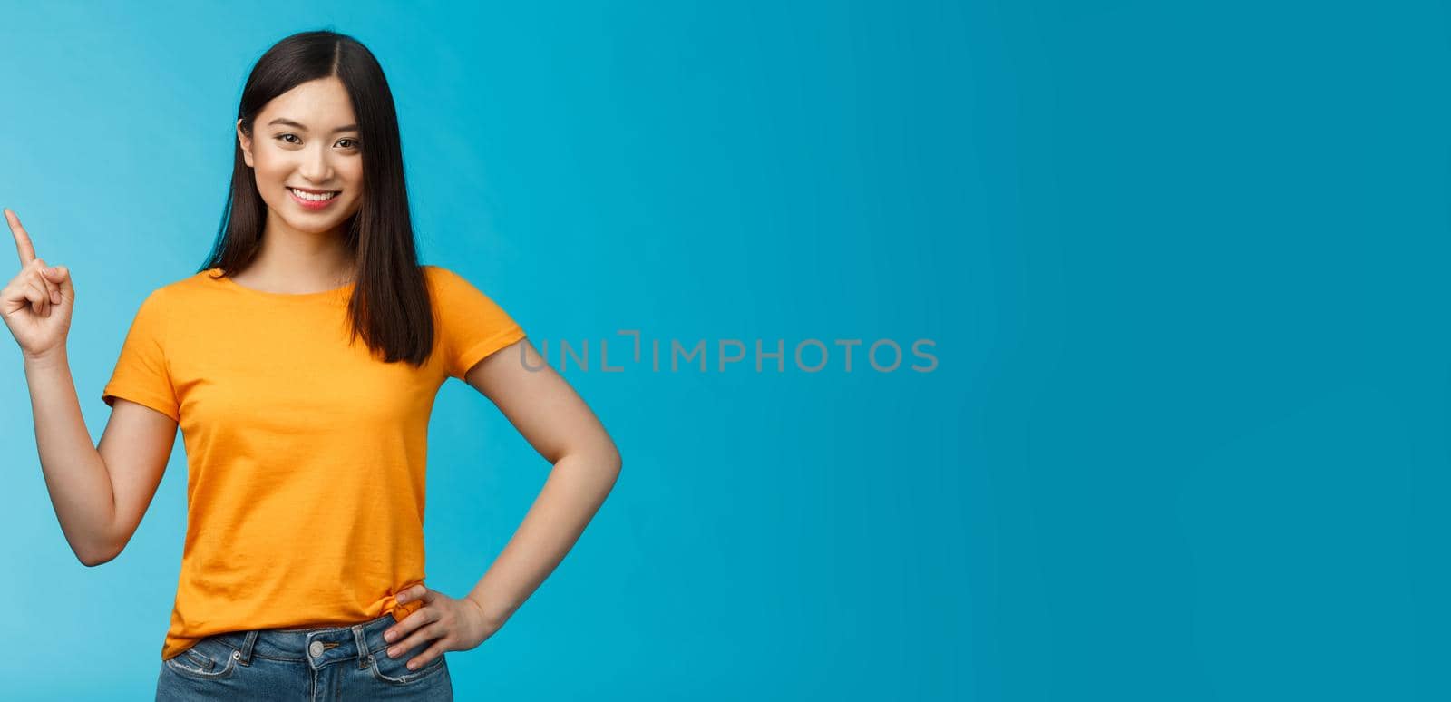 Happy confident asian girl self-assured pointing upper left corner, show way, smiling determined, assertive introduce online store promo, stand blue background in yellow t-shirt. Copy space