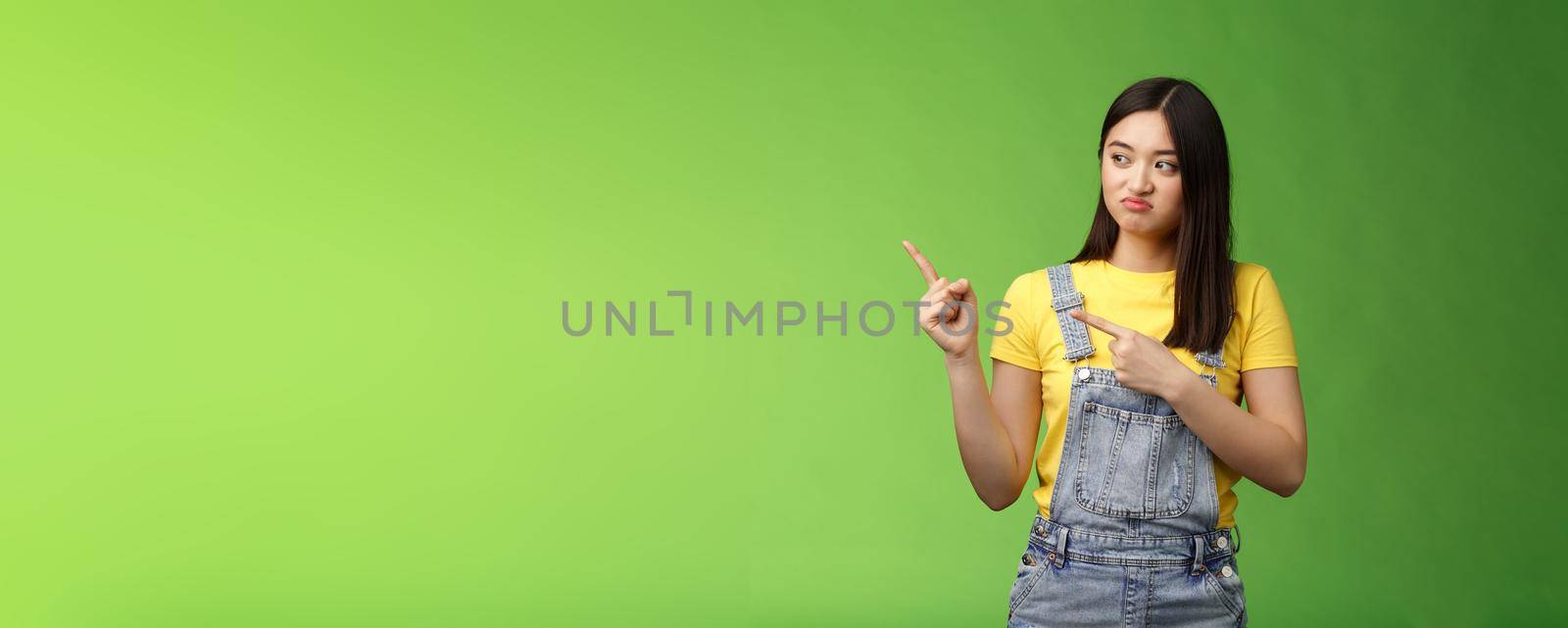 Upset cute asian female student pulling face regret shame, pity missing good chance buy product sale, pointing looking left disappointed, jealously gaze coworker, stand green background by Benzoix