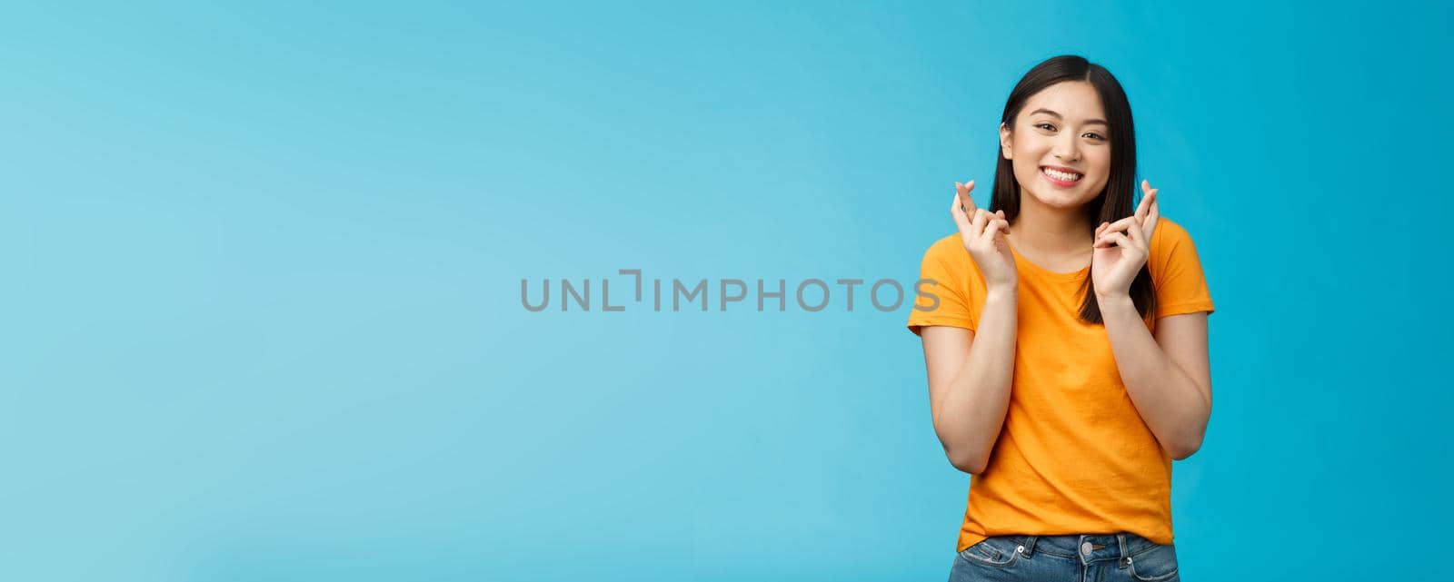 Cheerful smiling hopeful asian girl praying wish for good luck, cross fingers grinning toothy anticipate positive good news, stand blue background supplicating optimistic feelings hope win by Benzoix