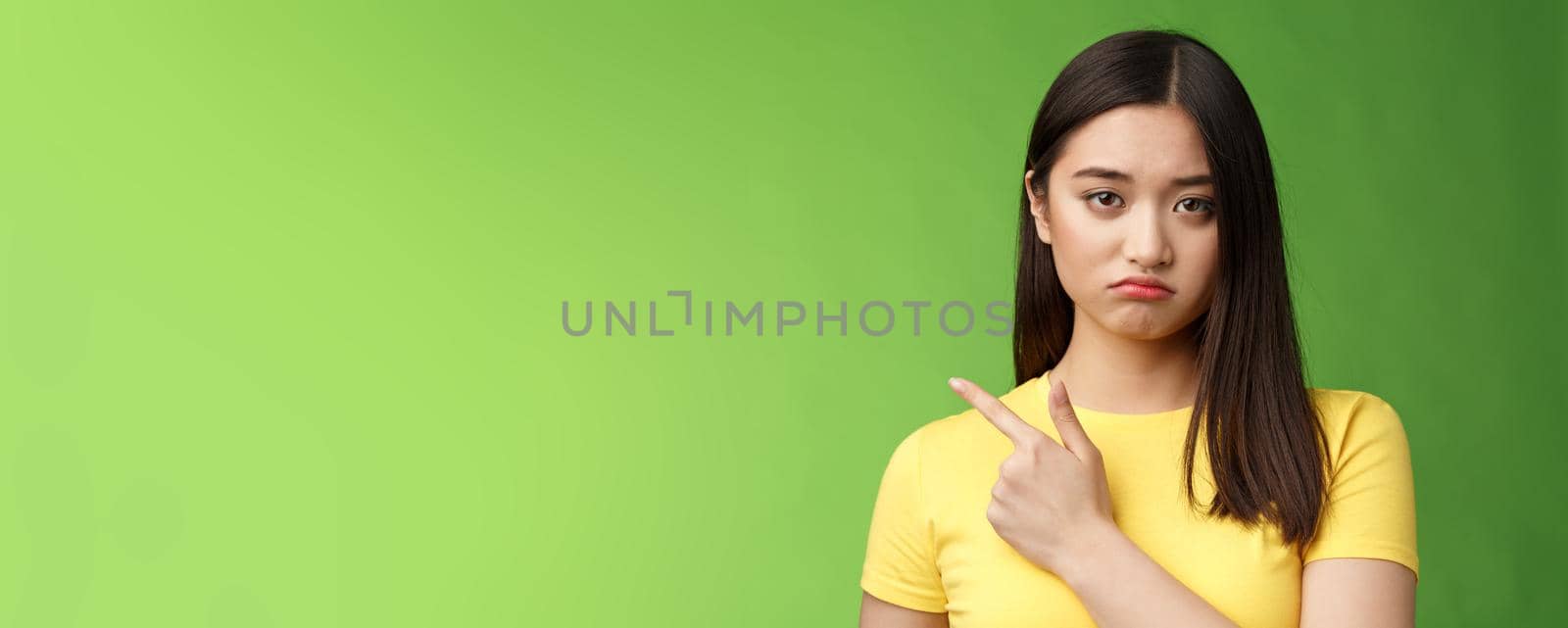 Grumpy upset cute asian girl complaining tired, drained during university exams, pointing unhappy, losing competition, pointing left, sulking displeased, stand green background reluctant by Benzoix