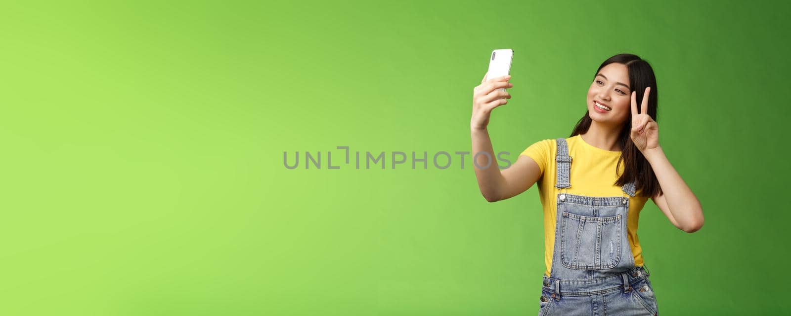 Tender cute asian brunette female, hold smartphone, posing selfie, look telephone screen, photographing, show victory peace sign, joyfully chatting friend abroad, stand green background. Copy space