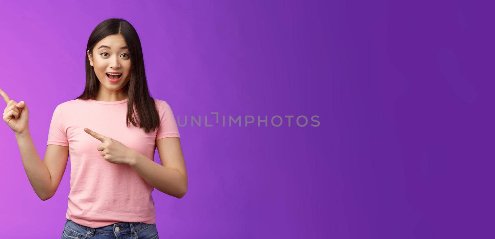 Enthusiastic surprised cute asian young girl showing promo lively grinning and telling about hot sales, pointing left indicating sideways, smiling joyfully camera, excited tell opening new store.