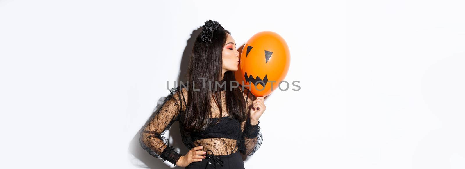 Stylish asian woman in gothic lace dress celebrating halloween, kissing orange balloon with face, standing over white background.