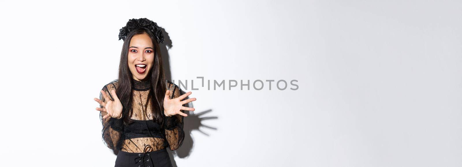 Image of excited asian woman in black gothic dress making big announcement, shaking hands and talking, standing over white background.