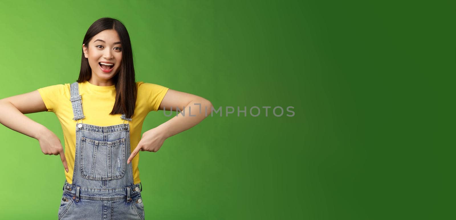 Lively energized asian cute female promote advertisement pointing fingers down, show you cool place, smiling camera joyful, telling great pleasant news stand green background emotional by Benzoix