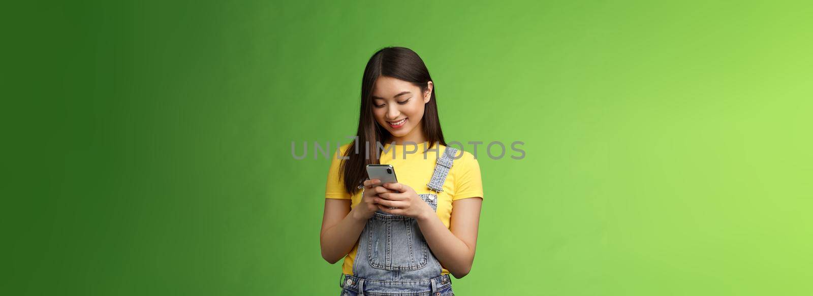 Outgoing cute asian brunette woman scroll online shop, purchase summer tour internet, hold smartphone, look amused telephone screen, use app, edit photos post social media, stand green background.