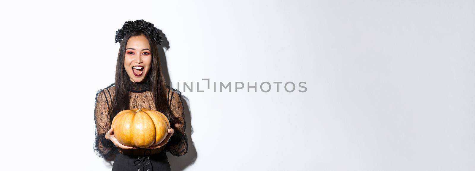 Image of joyful asian woman in witch costume, celebrating halloween, giving you pumpkin and looking scary, standing over white background.