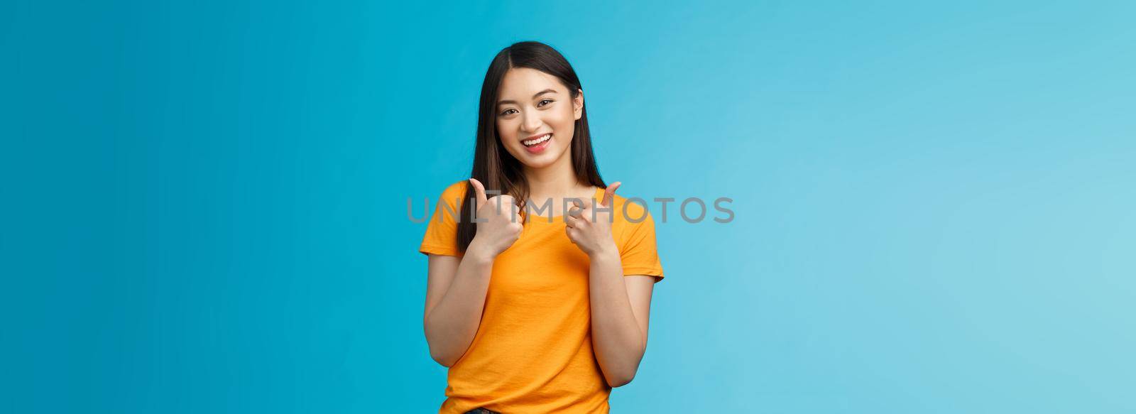 Supportive cute friendly asian girl give thumbs up and smiling, cheering for you, congratulate nice work, approving good idea, like nice outfit, grinning glad accept plan, stand blue background.