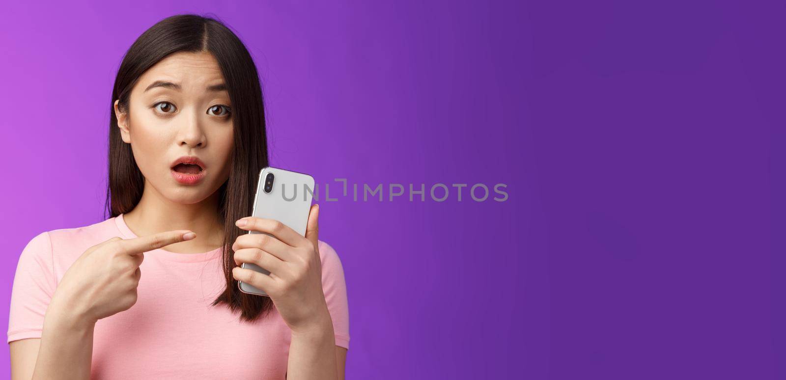 Concerned confused cute young asian woman hold smartphone open mouth, pointing finger telephone, look questioned upset, troubled see strange post online, stand purple background. Copy space