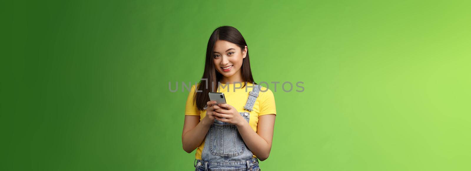 Cute tender asian gorgeous woman using smartphone, smiling carefree, look camera, texting friend, waiting boyfriend finish work, stand cafe green background, hold telephone, order food online by Benzoix