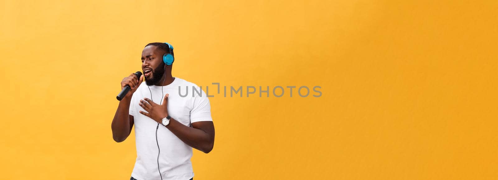Portrait of cheerful positive chic. handsome african man holding microphone and having headphones on head listening music singing song enjoying weekend vacation isolated on yellow background.