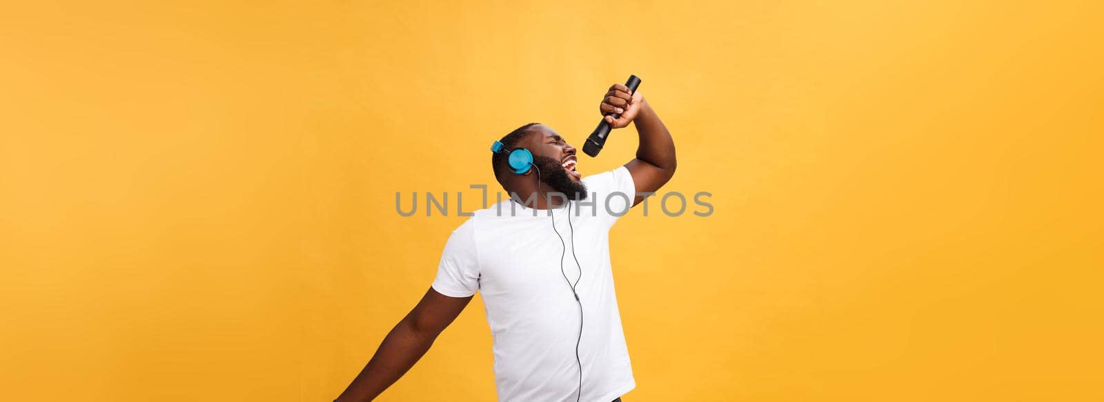 Portrait of cheerful positive chic. handsome african man holding microphone and having headphones on head listening music singing song enjoying weekend vacation isolated on yellow background.