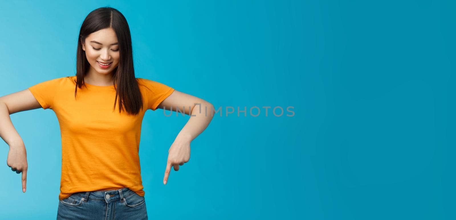 Cute tender feminine asian girl dark haircut look pointing down contemplating interesting product, stare curiously smiling amused, express enthusiasm interest try-out cool thing, blue background.