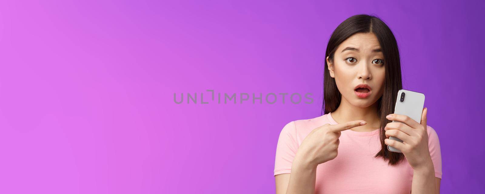 Concerned confused cute young asian woman hold smartphone open mouth, pointing finger telephone, look questioned upset, troubled see strange post online, stand purple background by Benzoix