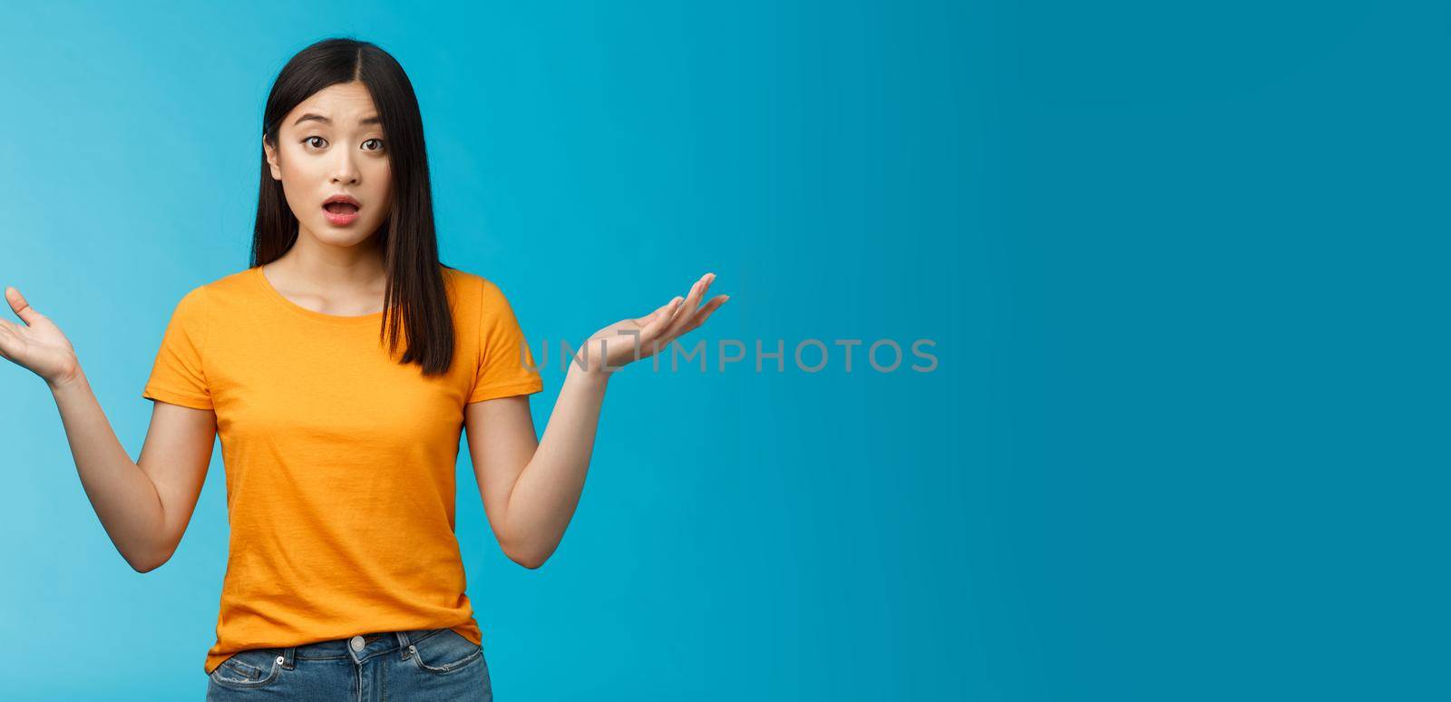 Questioned unaware young asian girl with dark short haircut shrugging hands spread sideways, look confused open mouth uncertain, being clueless, stand blue background wear yellow t-shirt by Benzoix