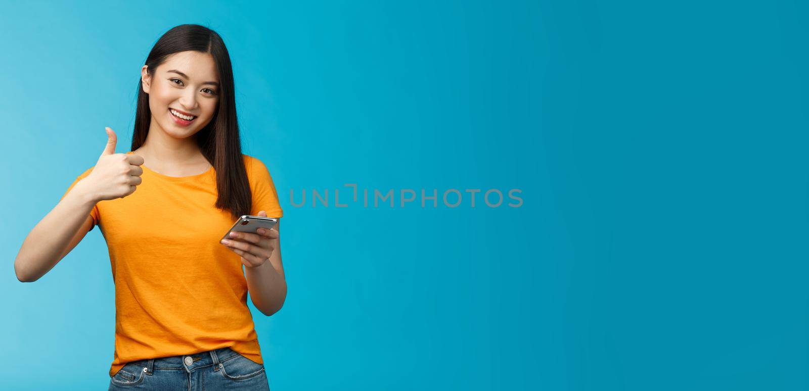 Satisfied cute asian brunette hold smartphone show thumb-up delighted, recommend awesome gadget, give approval good new app, agree friend posted good blog article, stand blue background by Benzoix