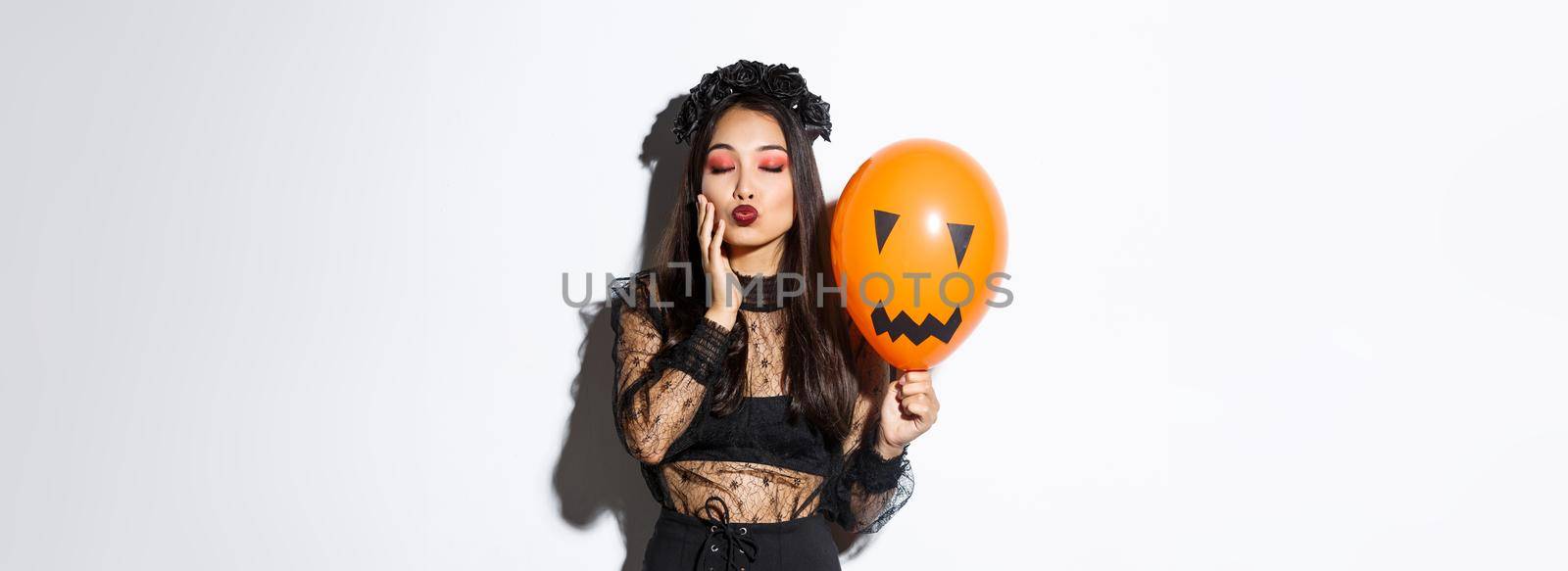 Image of beautiful stylish asian woman celebrating halloween, wearing witch costume, holding orange balloon with face.