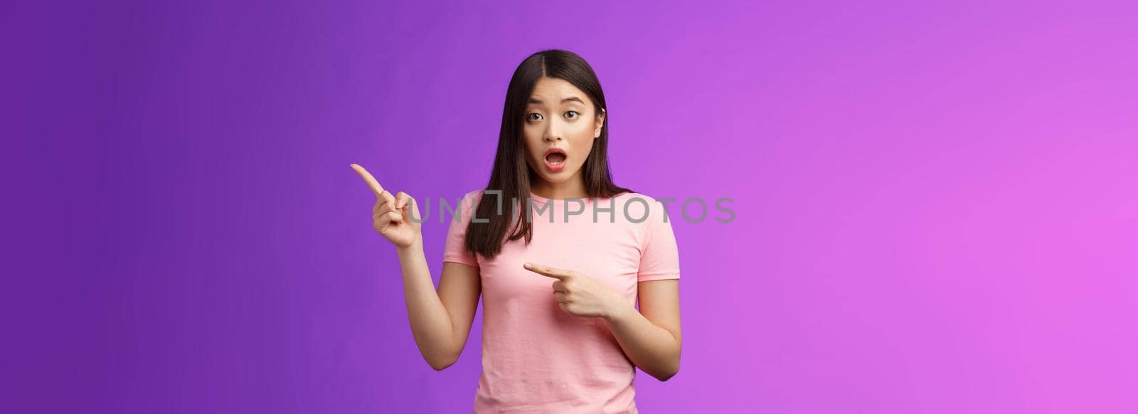 Excited alarmed cute asian female describe shocking scene pointing right, show index fingers sideways thrilling event, discuss amazing performance look camera worried, stand purple background.