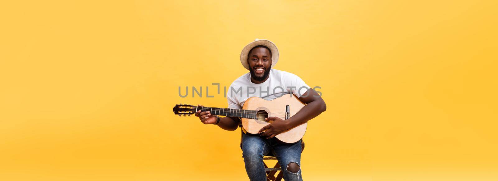 Muscular black man playing guitar, wearing jeans and white tank-top. Isolate over yellow background