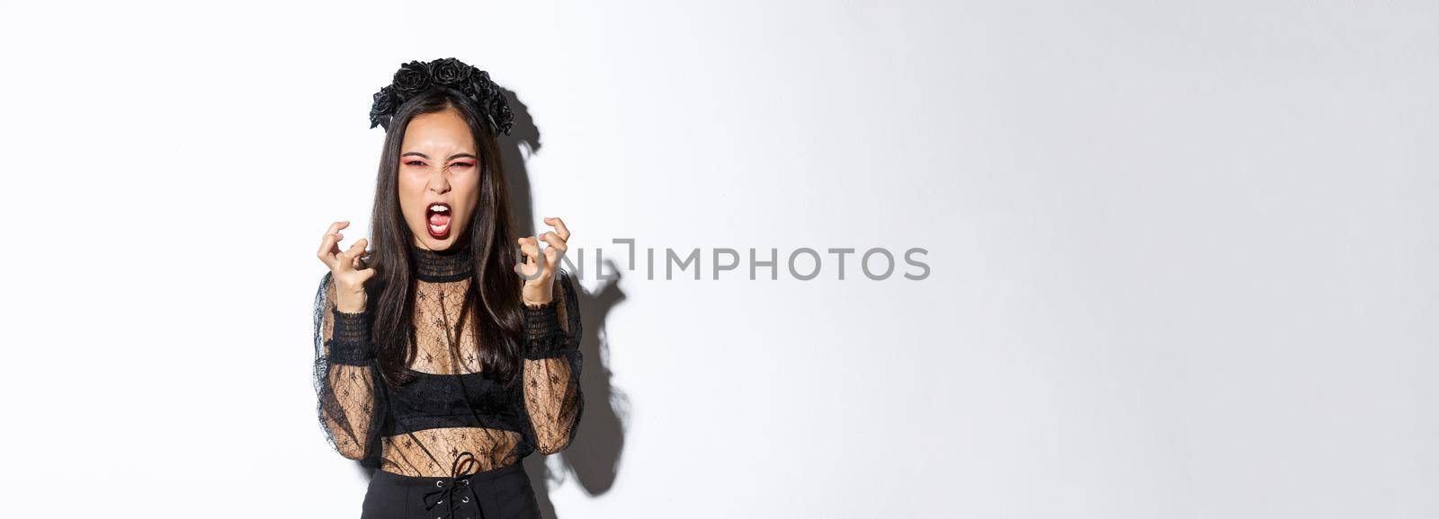 Image of mad woman in gothic lace dress and black wreath yelling angry and clenching fists, standing in halloween costume with furious expression, standing over white background by Benzoix