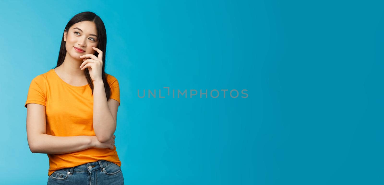 Joyful confident asian girl with dark haircut tilt head, dreaming, imaging interesting plan, smiling cunning, have idea, thinking and making choice, look up thoughtful, stand blue background.