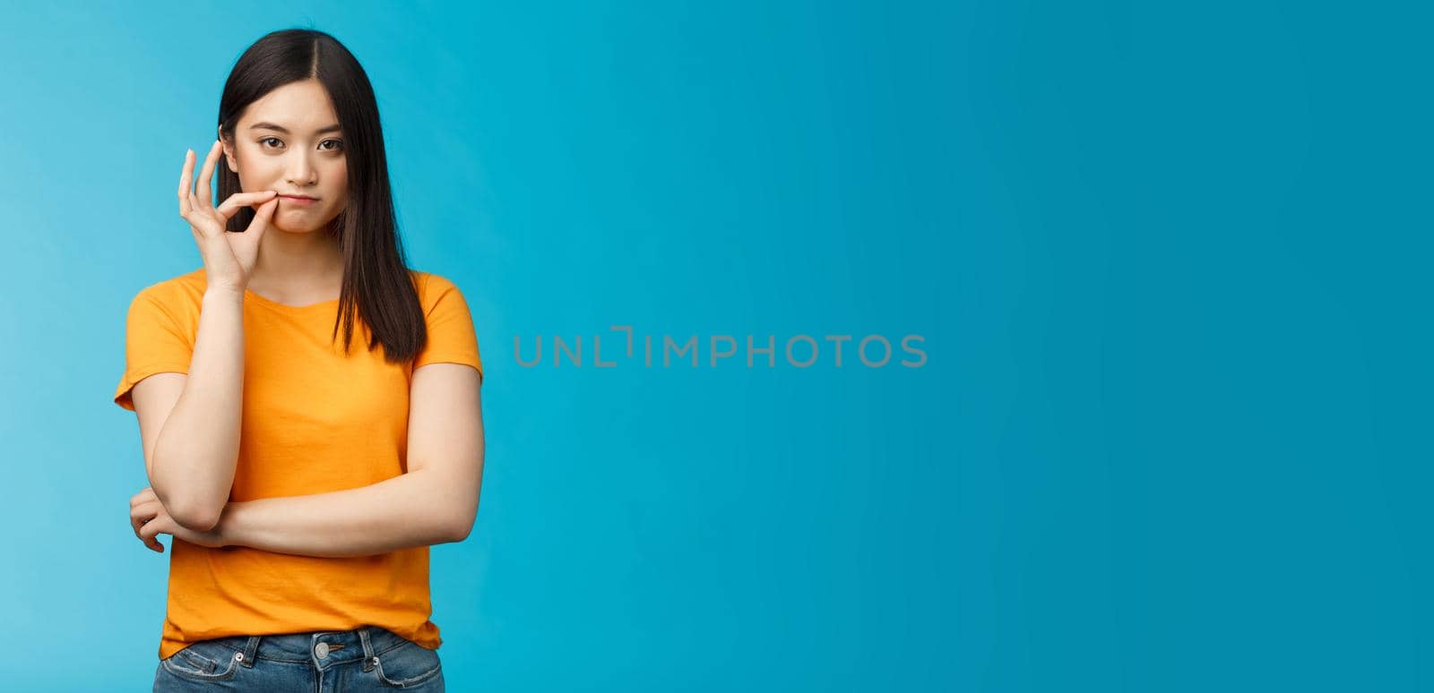 Serious-looking asian female friend promise keep secret, seal lips, hold zip near mouth look focused determined, stay silent and speechless, stand wearing yellow t-shirt blue background.