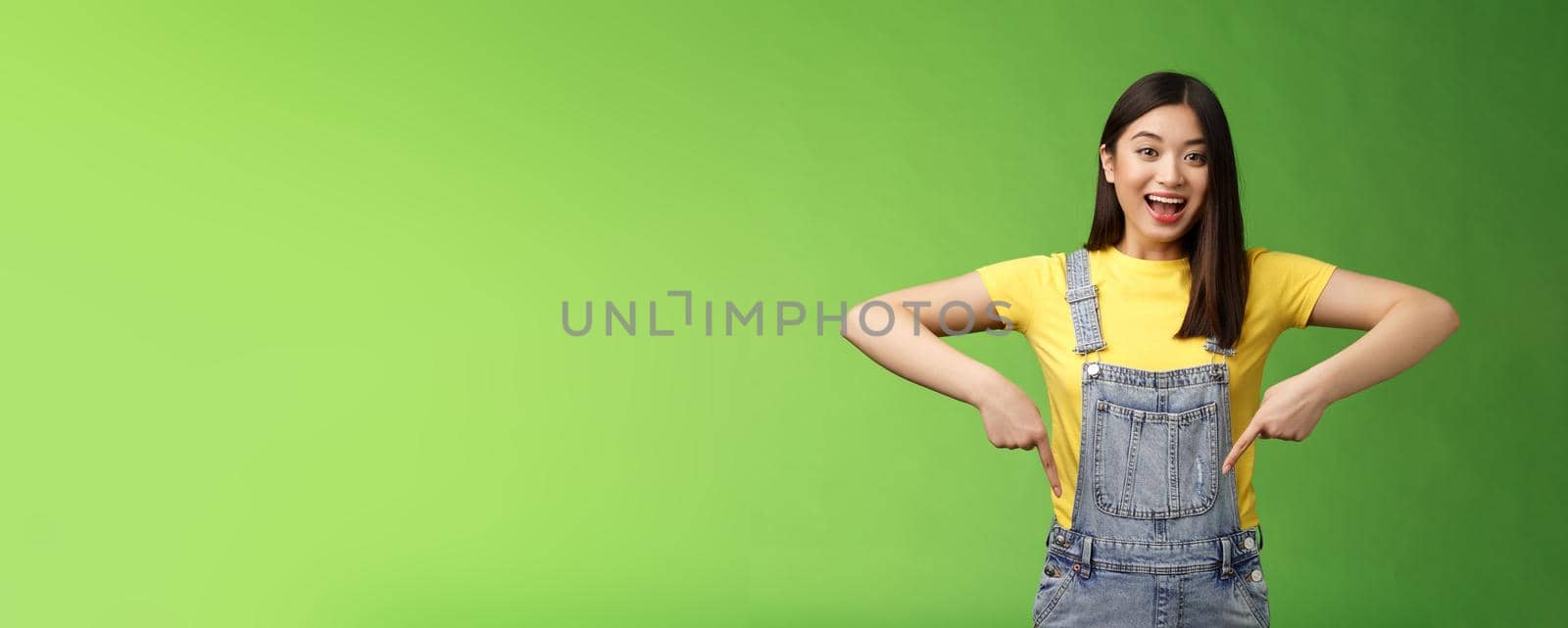 Lively energized asian cute female promote advertisement pointing fingers down, show you cool place, smiling camera joyful, telling great pleasant news stand green background emotional.