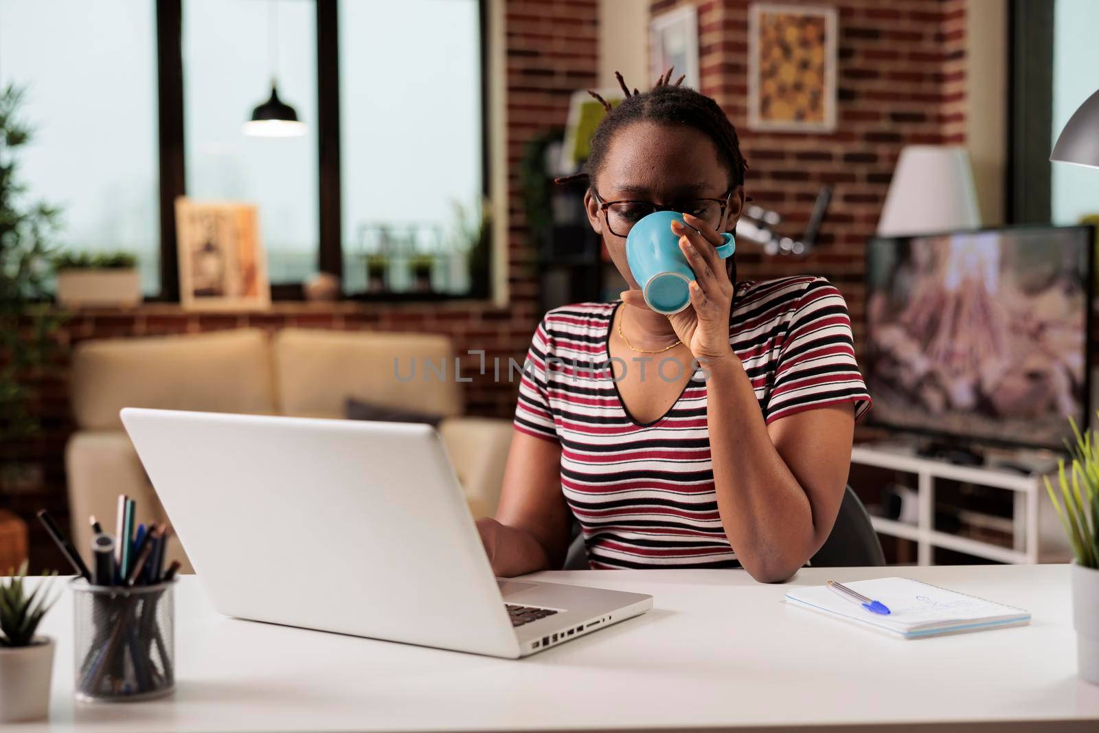 Remote student watching educational tutorial on laptop at home, drinking coffee. Woman attending online courses class, young freelancer working on internet, holding tea mug