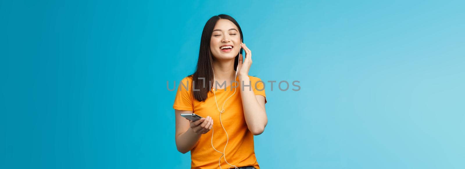 Lovely carefree cute asian girl enjoy awesome music boost mood listening favorite songs, touch earphone close eyes from pleasure delight, singing along, hold smartphone stand blue background.