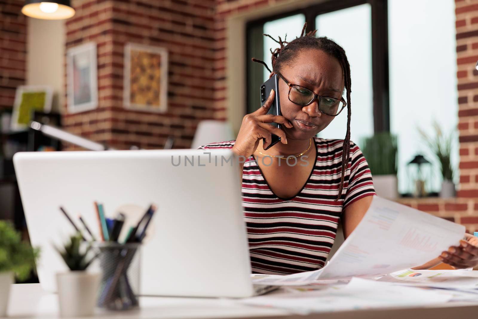Serious freelancer answering mobile phone call, discussing report, holding documents, looking at research statistics. Employee working from home, remote worker talking on smartphone