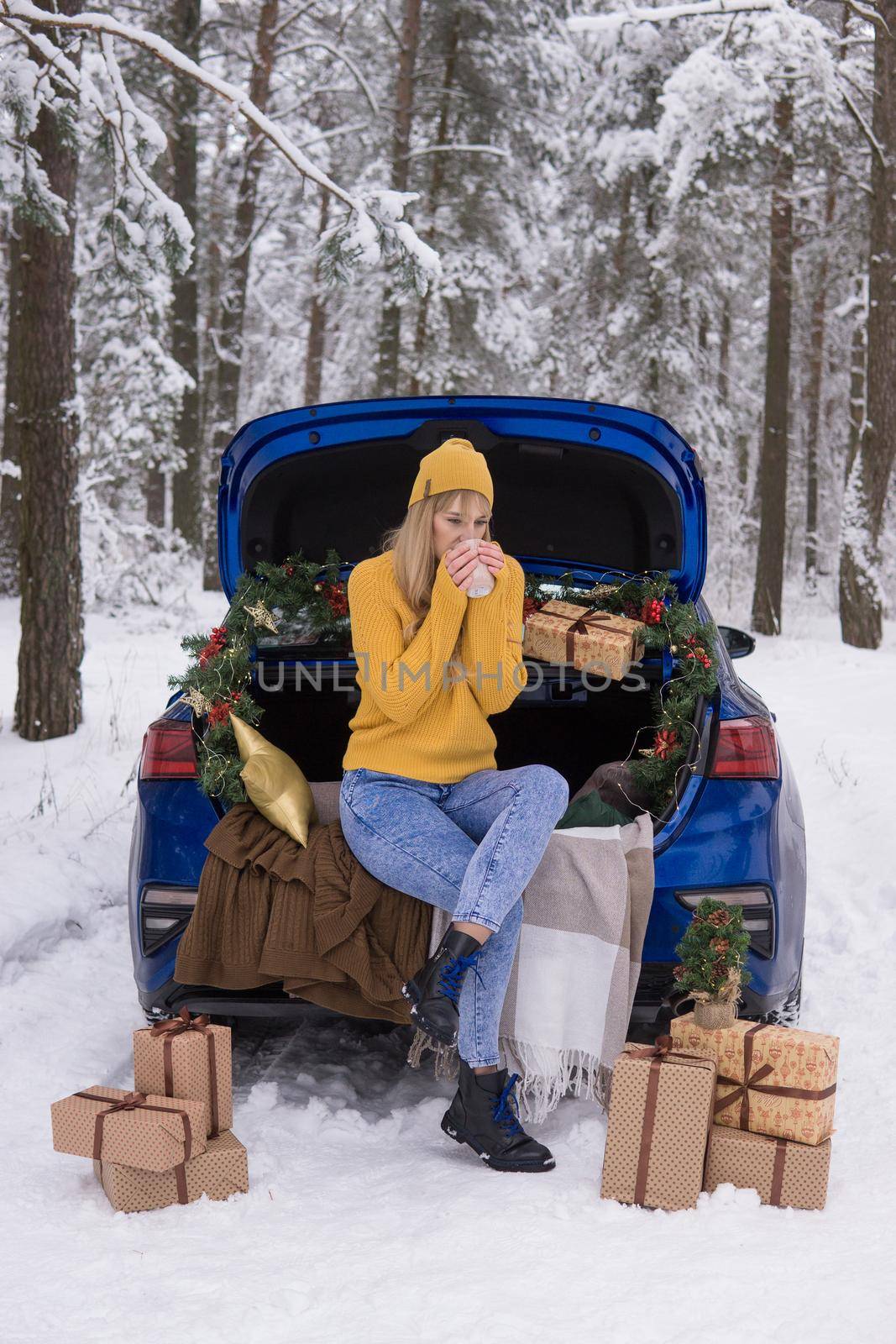 A woman in a winter snow-covered forest in the trunk of a blue car decorated with Christmas decor. The concept of Christmas and winter holidays.