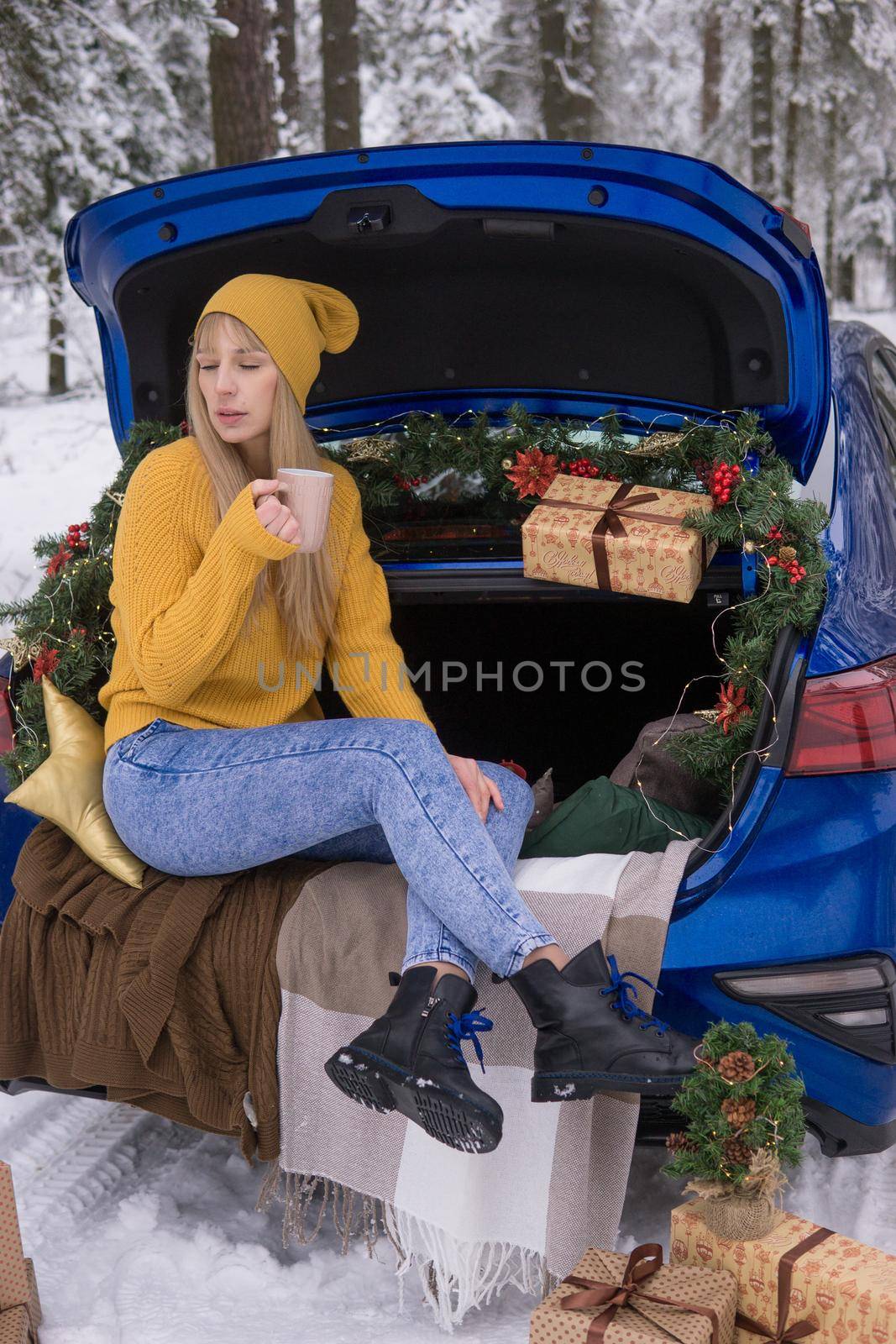 A woman in a winter snow-covered forest in the trunk of a blue car decorated with Christmas decor. The concept of Christmas and winter holidays.